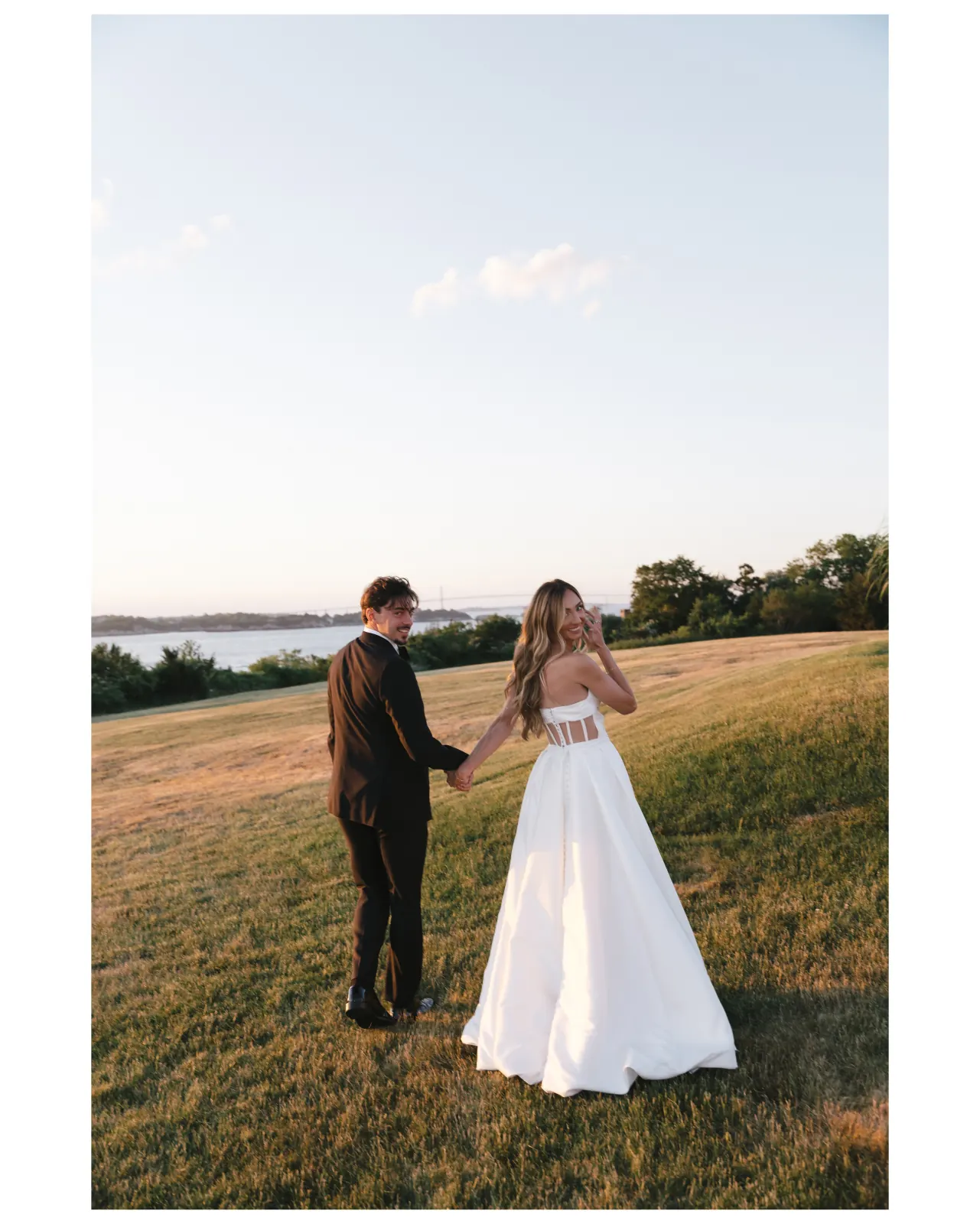 Caite and Sean hold hands on the lawn with Narragansett Bay behind them at golden hour