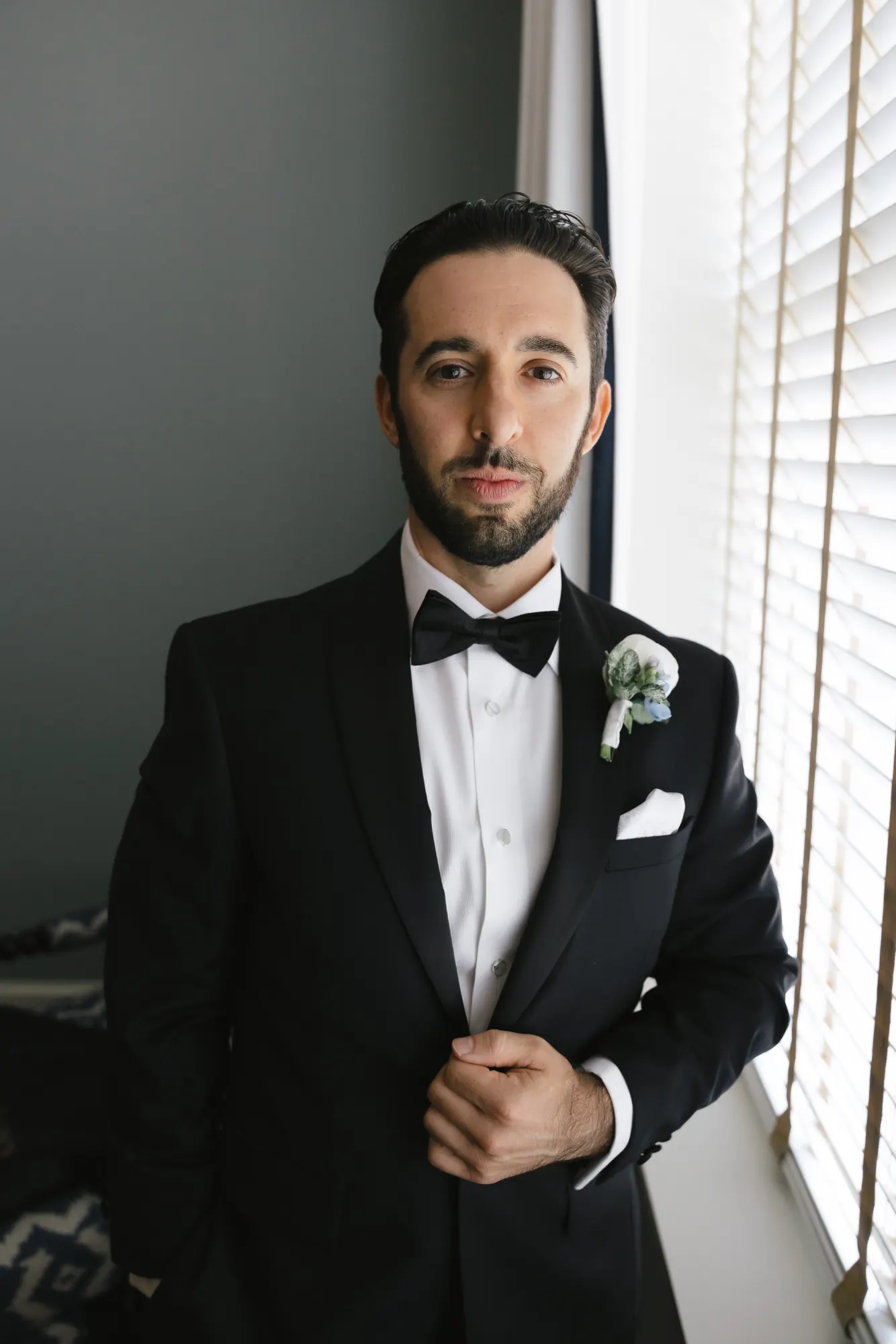 Groom in a black tuxedo and white boutonniere by the window light