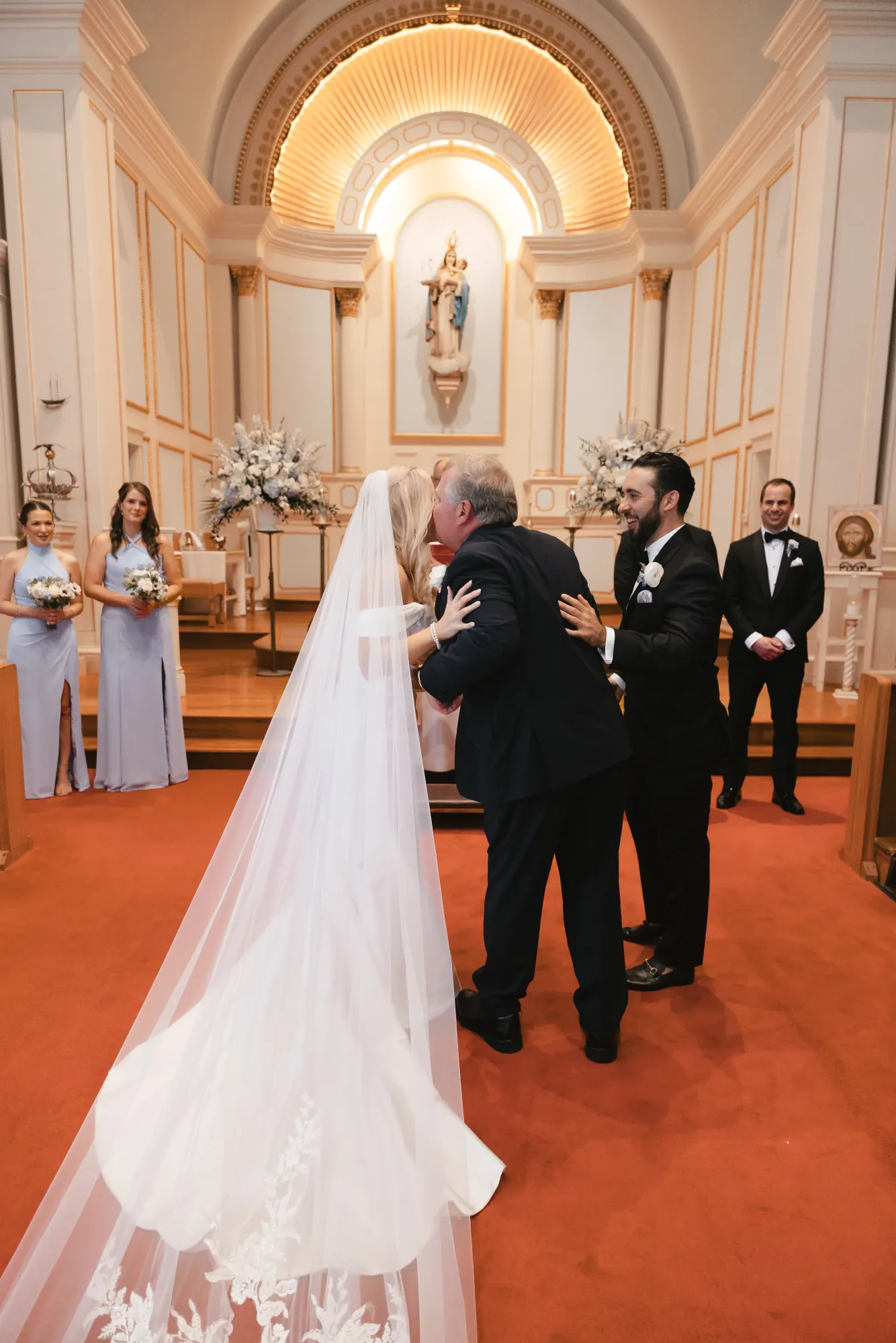 The bride's father gives her away at the front of the church, her lace veil trailing