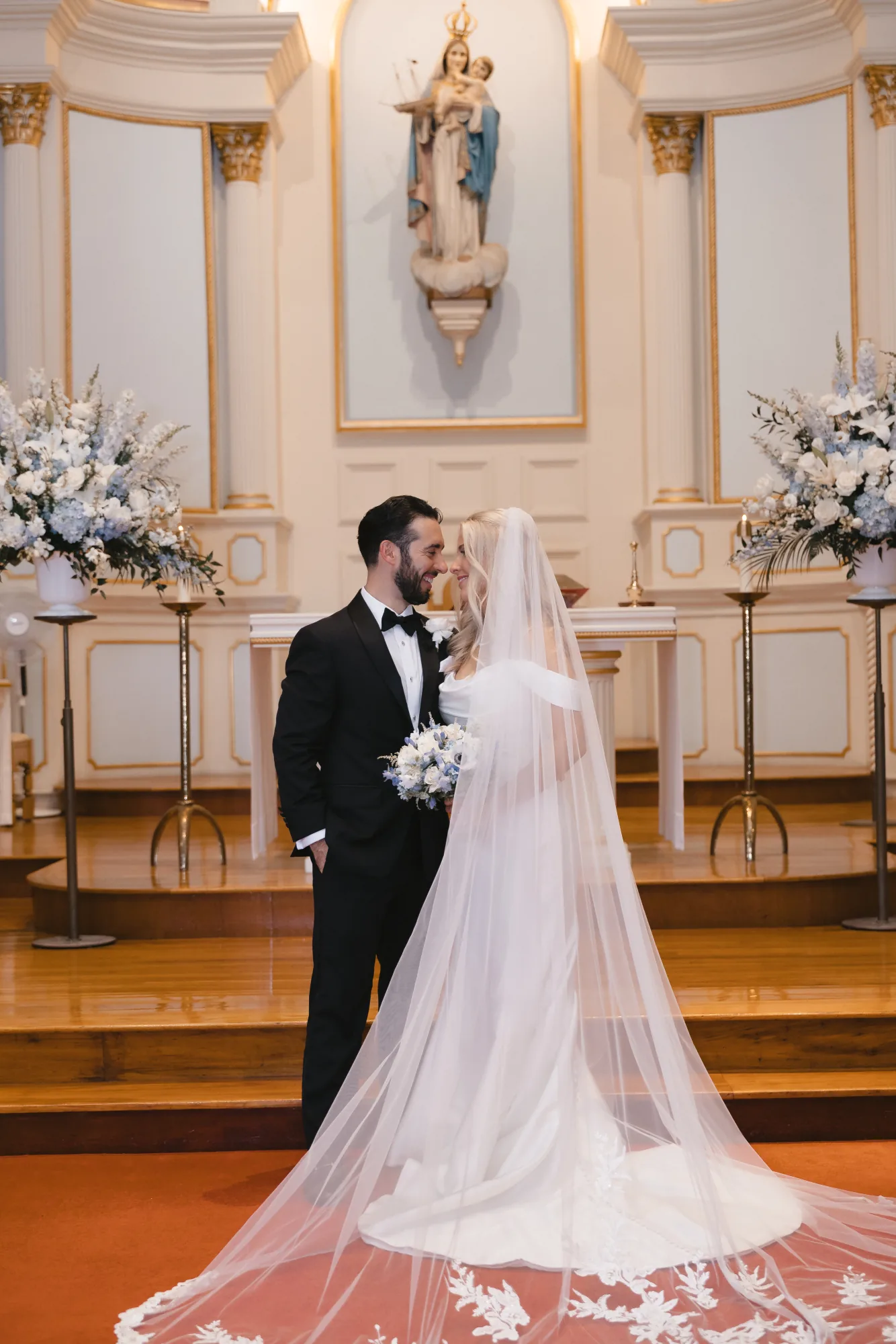 Bride and groom smile at each other at the altar of Our Lady of Good Voyage in Gloucester, Massachusetts, framed by blue and white florals