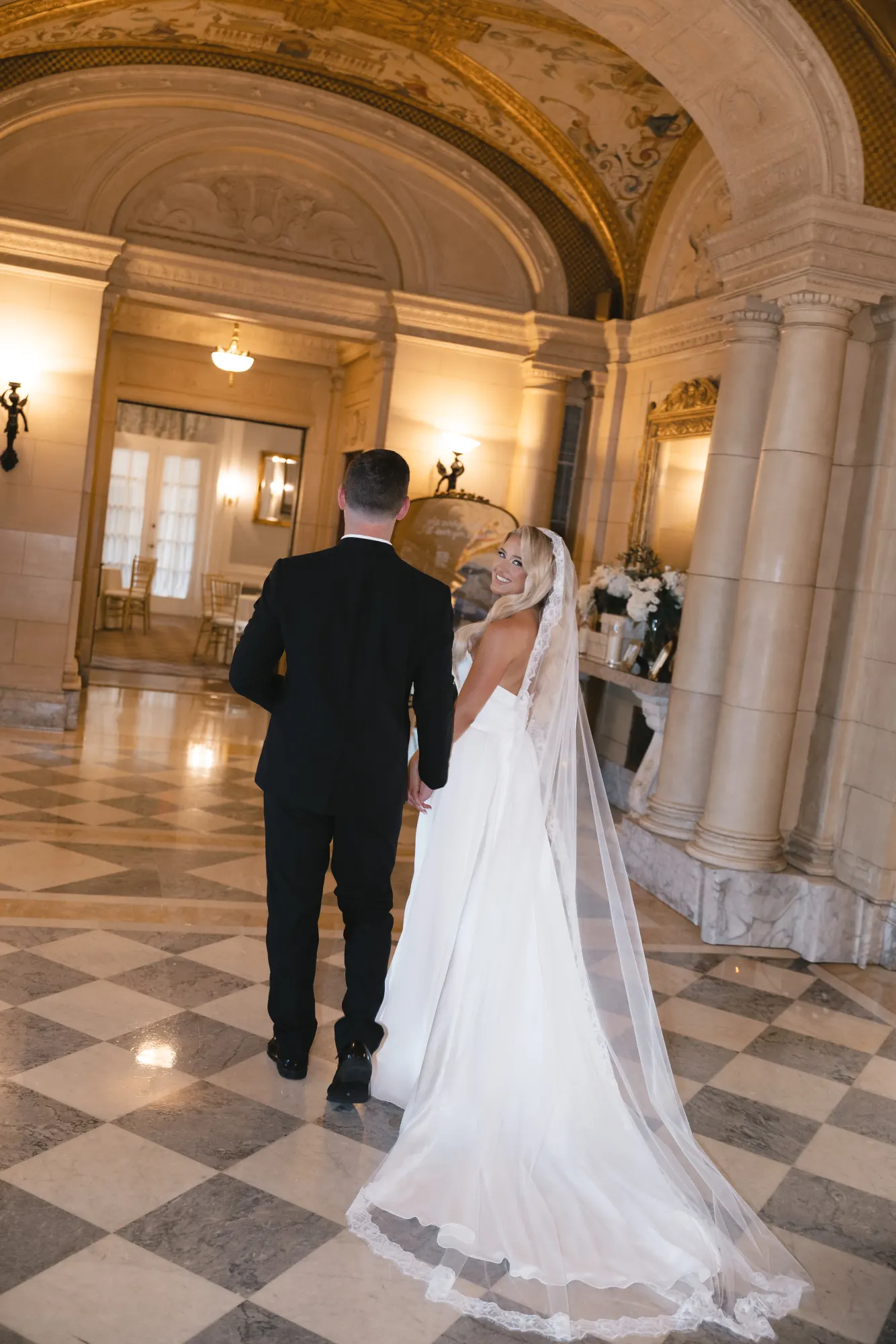 The couple crosses the marble checkerboard hall hand in hand