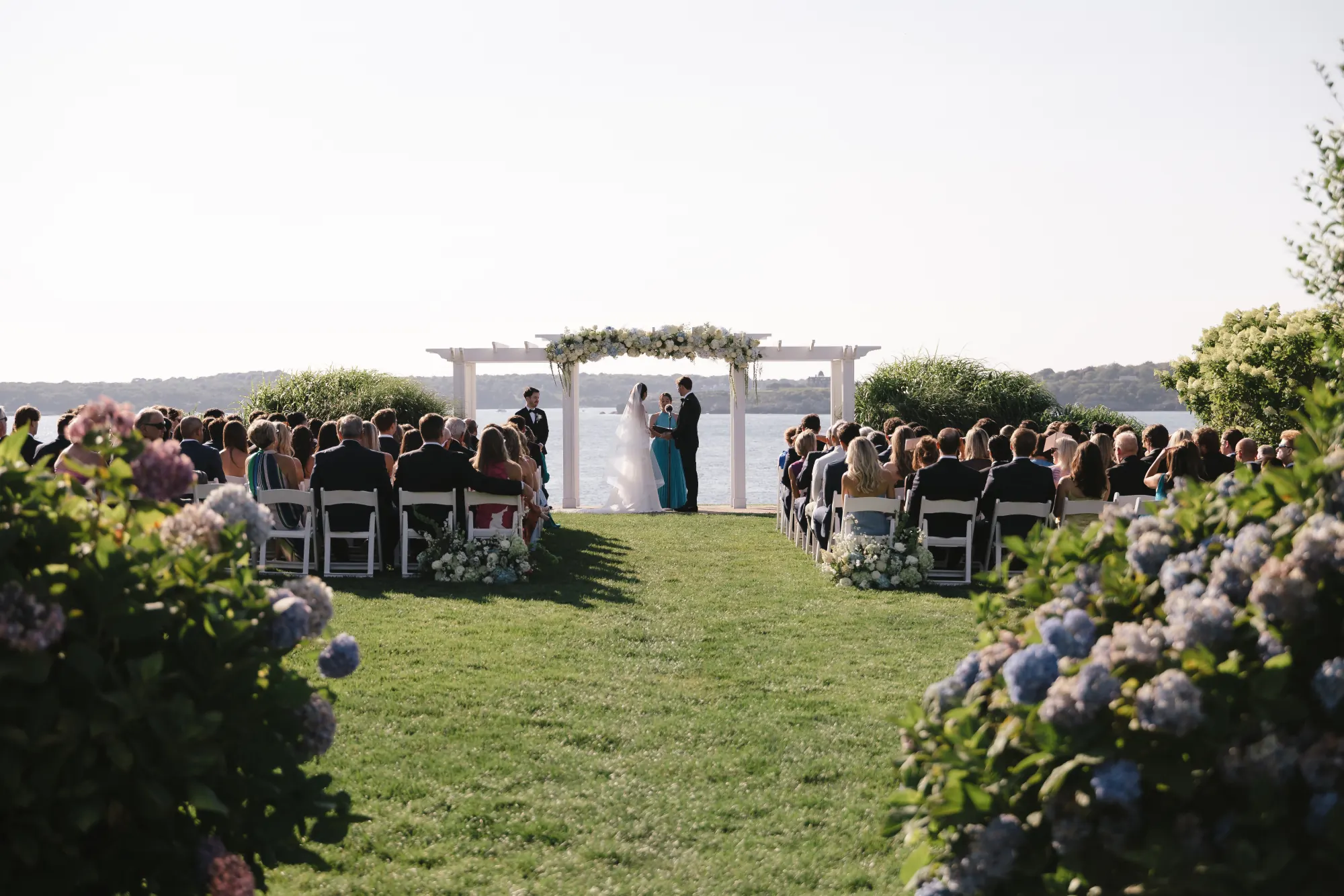 Wide view of the Bridge Lawn ceremony over Narragansett Bay, hydrangeas in bloom