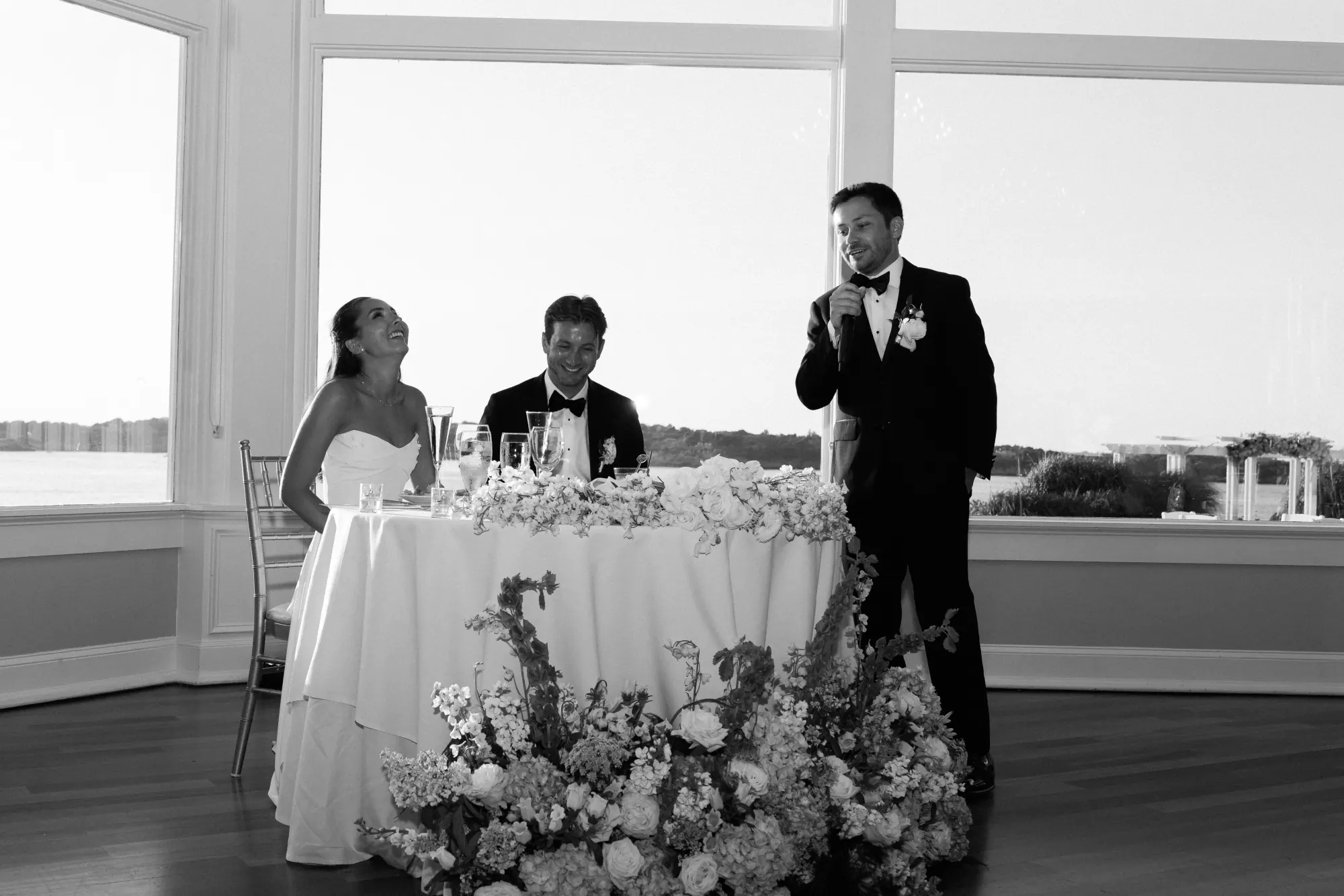 Black and white of the toasts at the flower-wrapped sweetheart table