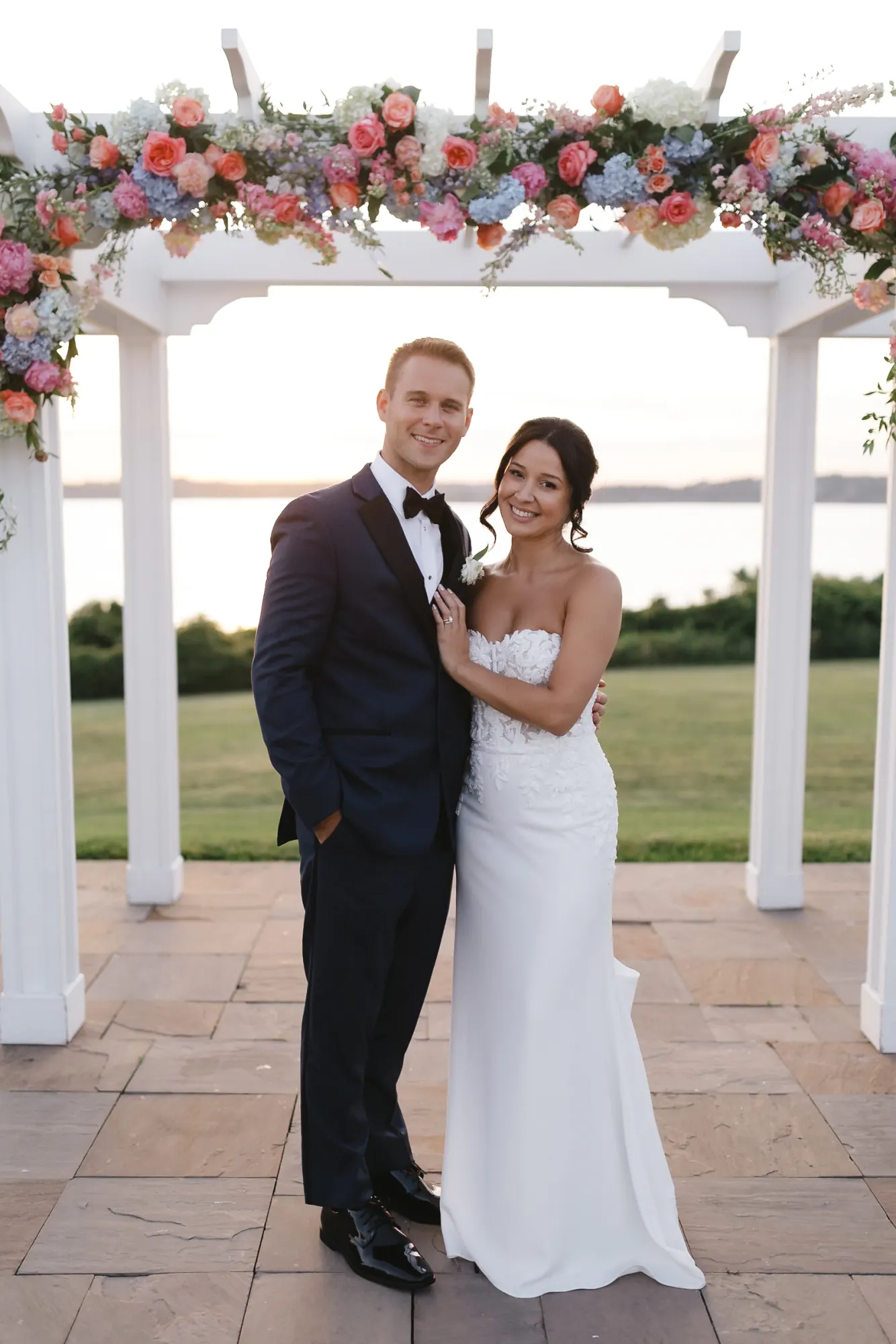 Bride and groom under a floral arbor with Narragansett Bay behind at golden hour, OceanCliff Newport