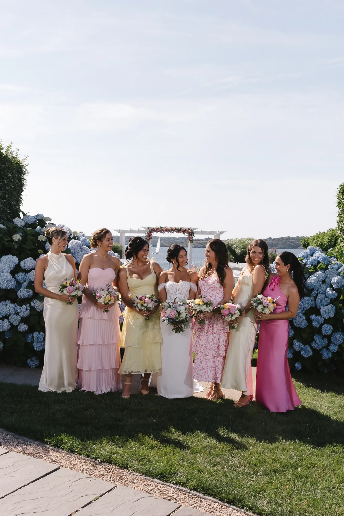 Bride with her bridesmaids in pastel dresses holding bouquets by the blue hydrangeas and the bayfront arbor at OceanCliff in Newport, Rhode Island